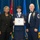 Airman Leadership School distinguished graduate A United States Air Force graduation ceremony at the Airman Leadership School, where a female airman is receiving her diploma. She is flanked by a civilian woman in black attire on the left and a senior enlisted military officer in dress uniform on the right. The setting features multiple flags including the U.S. flag and what appears to be the Jordanian flag.