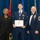Airman Leadership School distinguished graduate A formal military ceremony photograph showing three individuals posing together, with the central figure holding a framed diploma or certificate. The setting appears to be a United States Air Force Airman Leadership School graduation ceremony, indicated by the visible certificate header and the medal worn by the recipient. Two presenters flank the graduate — a civilian woman in a black suit on the left and a senior non-commissioned officer in dress uniform on the right.