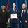 Airman Leadership School distinguished graduate A formal military graduation or awards ceremony featuring three individuals posing for a photo. The central figure, a young airman in U.S. Air Force dress blues, is holding a framed diploma and wearing a medal around his neck. He is flanked by a civilian woman in a black suit on the left and a senior non-commissioned officer (Chief Master Sergeant) in dress blues with multiple medals on the right.