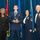 Airman Leadership School Academic Achievement Award A formal military awards ceremony featuring four individuals posing for a photo. A young airman in the center is holding a golden eagle trophy and wearing a medal around his neck, flanked by two senior non-commissioned officers in dress uniforms and a civilian woman in a black suit. The backdrop includes a US flag, what appears to be a Jordanian flag, and Air Force flags against a blue curtain.