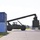 Fort McCoy supports USTRANSCOM's TMS prototype operations A military-green reach stacker (container handler) is actively lifting or moving a green shipping container in what appears to be a military logistics yard. Blue and green ISO shipping containers are stacked on the left side, with additional containers and structures visible in the background. The scene depicts a container handling operation at what appears to be a military installation or depot.