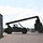 Fort McCoy supports USTRANSCOM's TMS prototype operations A large reach stacker or container handler is operating in what appears to be a military logistics yard, maneuvering shipping containers. The vehicle is captured in silhouette against a bright sky, with its extended boom arm holding or positioning a container on the right side. The facility contains multiple stacked shipping containers in blue and olive/military green colors.