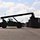 Fort McCoy supports USTRANSCOM's TMS prototype operations A large reach stacker or container handler vehicle is silhouetted against a partly cloudy sky, operating in what appears to be a military or industrial storage yard. The vehicle's extended boom arm is positioned over a row of stacked shipping containers on the right side of the image. The strong backlighting creates a dramatic silhouette effect, obscuring fine details of the machinery.