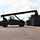 Fort McCoy supports USTRANSCOM's TMS prototype operations A large reach stacker or container handler vehicle is silhouetted against a bright sky on a paved surface, appearing to be at a military installation or logistics yard. The heavy equipment is positioned near a row of military-style shipping containers or equipment cases. A single operator is visible in the cab of the vehicle.