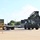 Fort McCoy supports USTRANSCOM's TMS prototype operations A military-green reach stacker or container handler is positioned in a paved logistics yard, facing a flatbed trailer loaded with a green shipping container. The scene appears to be a military or government logistics facility, indicated by the olive-drab color scheme of both the equipment and container. The operation suggests an active container loading or unloading operation.