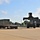 Fort McCoy supports USTRANSCOM's TMS prototype operations A military-style heavy forklift (likely a Rough Terrain Container Handler) is positioned near a flatbed semi-trailer carrying a large olive drab military shipping container in an open logistics yard. The scene appears to be a military installation or depot, with a large industrial building visible in the background. The forklift appears to be preparing to load or unload the container from the trailer.