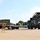 Fort McCoy supports USTRANSCOM's TMS prototype operations A military or industrial logistics yard showing a large olive-drab reach stacker (container handler) positioned near a flatbed semi-trailer carrying a green shipping container. The scene appears to depict an active container loading or unloading operation at what looks like a military installation or freight depot. Yellow safety goal-post structures are visible in the foreground, likely used as height clearance indicators.