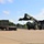 Fort McCoy supports USTRANSCOM's TMS prototype operations A military-green reach stacker (container handler) is positioned to lift or place a large olive-drab shipping container onto a flatbed semi-truck trailer in an open paved yard. The equipment and container appear to be military in nature based on their coloring. At least one person is visible near the cab of the truck, likely supervising or guiding the operation.
