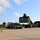 Fort McCoy supports USTRANSCOM's TMS prototype operations A military or government logistics facility where a reach stacker (container handler) is lifting an olive drab/military green shipping container, apparently positioning it onto or removing it from a flatbed semi-truck trailer. The scene takes place on a large concrete apron adjacent to a warehouse building, with stacked intermodal containers visible on the left side. The equipment and container colors suggest a U.S. military installation.