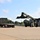 Fort McCoy supports USTRANSCOM's TMS prototype operations A military-painted olive green reach stacker (container handler) is positioned to lift or place a shipping container onto a flatbed semi-truck trailer in a paved logistics yard. The operation is taking place adjacent to a dark-colored industrial or military building. The scene suggests a container loading or unloading operation at a military or government facility.