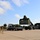 Fort McCoy supports USTRANSCOM's TMS prototype operations A military logistics operation is underway at what appears to be a military installation or depot. A large olive-drab reach stacker (container handler) is lifting a military shipping container onto or off of a flatbed semi-truck trailer. Stacks of similar military containers are visible to the left, alongside a warehouse-style building on the right.