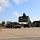 Fort McCoy supports USTRANSCOM's TMS prototype operations A military reach stacker (painted olive drab/green) is lifting a dark green military container at what appears to be a military logistics or cargo handling facility. A white civilian flatbed semi-truck is positioned nearby, likely to receive or has just delivered the container. The facility includes a concrete apron, storage containers along the left side, and a dark-colored warehouse building on the right.