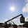 Fort McCoy supports USTRANSCOM's TMS prototype operations A large reach stacker or telescopic handler is lifting a shipping container at what appears to be a military or industrial logistics facility. The image is taken from a low angle against a bright sun, creating a dramatic silhouette effect with visible lens flare. The vehicle's extended boom arm holds a container suspended in the air while an operator is visible in the cab.