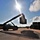 Fort McCoy supports USTRANSCOM's TMS prototype operations A reach stacker or container handler is lifting a large shipping container in what appears to be a logistics yard or military installation. The image is taken against a bright midday sun, creating strong silhouettes and dramatic lens flare. The facility features multiple storage containers and light poles arranged along a paved yard.