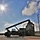 Fort McCoy supports USTRANSCOM's TMS prototype operations A large reach stacker or container handler is silhouetted against a bright midday sky, actively lifting or positioning a shipping container in what appears to be a logistics or port facility. The image is taken from a low angle with strong backlighting from the sun, creating dramatic silhouette effects. The facility includes security fencing, utility poles, and additional shipping containers in the background.