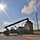 Fort McCoy supports USTRANSCOM's TMS prototype operations A reach stacker or container handler is actively lifting or positioning a large shipping container in what appears to be a military or industrial logistics yard. The image is taken against a bright sun, creating strong silhouette effects and lens flare. The facility appears secured with fencing and utility poles visible in the background.