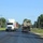Fort McCoy supports USTRANSCOM's TMS prototype operations A two-lane road scene showing oncoming traffic including a white International semi-truck with a box trailer and a flatbed trailer transporting what appears to be a military vehicle painted in tan/olive drab colors. The setting appears to be a suburban or semi-rural area with well-maintained grass, utility poles, and mature trees lining both sides of the road. A person on a riding lawn mower is visible on the left shoulder of the road.