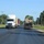 Fort McCoy supports USTRANSCOM's TMS prototype operations A two-lane road scene showing a convoy of vehicles including a white International semi-truck with a box trailer and a flatbed trailer transporting what appears to be a military tactical vehicle (likely an MRAP or similar armored vehicle). The setting appears to be a military installation or base, suggested by the well-maintained grounds, flag visible in the background, and the military equipment being transported. The image was taken during daytime under clear blue skies in a summer or late spring setting.