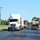 Fort McCoy supports USTRANSCOM's TMS prototype operations A convoy of semi-trucks travels along a two-lane road through what appears to be a military installation or rural area with well-maintained grass. The lead vehicle is a white International semi-truck with a flatbed trailer, followed by a maroon/yellow truck, and a flatbed trailer on the right carrying damaged or decommissioned military vehicles. A person on a riding lawn mower is visible on the left side of the road.