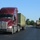 Fort McCoy supports USTRANSCOM's TMS prototype operations A dark red Kenworth semi-truck is traveling on a two-lane road hauling a flatbed trailer loaded with an olive-green military shipping container marked 'US/HCC4817'. In the opposing lane, a second flatbed truck is transporting what appears to be a military wheeled vehicle. The setting appears to be a military installation or base, suggested by the manicured grounds, wide roads, and military equipment being transported.