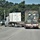 Fort McCoy supports USTRANSCOM's TMS prototype operations Two semi-trucks are traveling on a road near what appears to be a military installation or checkpoint. The lead truck carries a white shipping container, while the second truck transports a military-style camouflage container secured with yellow straps. Road signage including a STOP sign, pedestrian crossing sign, and an electronic message board displaying '234' are visible in the scene.
