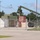 Fort McCoy supports USTRANSCOM's TMS prototype operations The image shows a military logistics or staging area enclosed by a chain-link fence, featuring shipping containers in various colors and a large olive-drab military crane (Kalmar brand) with its boom extended. Personnel in military uniform and civilian clothing are visible within the compound. The setting appears to be a military installation or base, likely during a loading or equipment staging operation.