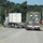 Fort McCoy supports USTRANSCOM's TMS prototype operations Two flatbed semi-trucks are traveling on a road, each carrying large cargo containers. The truck in the foreground carries a military-style camouflage-painted container, while the truck behind carries a white intermodal shipping container. The scene appears to be near a controlled intersection or installation entrance, indicated by road signs and an electronic message board.