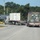 Fort McCoy supports USTRANSCOM's TMS prototype operations Two flatbed semi-trucks are traveling on a two-lane road, each carrying large shipping containers. The truck on the right carries a military-style olive drab/camouflage container marked with 'USAU' markings, while the left truck carries a white civilian shipping container. The scene includes road signage including a stop sign, a diamond warning sign, and an electronic message board displaying '123'.