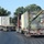 Fort McCoy supports USTRANSCOM's TMS prototype operations Two flatbed semi-trucks are traveling on a paved road, each carrying large cargo containers. The lead truck carries a military-style camouflage-painted container with visible weight markings, while the second truck carries a white shipping container. The scene appears to be in a rural or semi-rural area with trees and utility poles visible in the background.