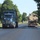 Fort McCoy supports USTRANSCOM's TMS prototype operations Two large trucks are traveling on a tree-lined road, likely on or near a military installation based on the visible flag and signage. The left truck is a blue Kenworth semi-truck hauling a green container, while the right truck is a flatbed hauling what appears to be a damaged or worn military tactical vehicle (MRAP or similar). The setting features well-maintained grass lawns and mature trees consistent with a military base environment.