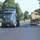Fort McCoy supports USTRANSCOM's TMS prototype operations Two large trucks are traveling on a paved road lined with mature trees and green grass. The left vehicle is a blue Kenworth semi-truck hauling what appears to be a green cargo container, while the right vehicle is a flatbed trailer transporting what appears to be a military vehicle (likely a tan/beige armored or tactical vehicle). The setting appears to be a military installation or base, suggested by the manicured grounds, flagpole, and nature of the cargo.
