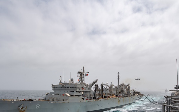 A replenishment-at-sea (RAS) operation is underway between two naval vessels in open ocean conditions. A gray auxiliary/replenishment ship bearing hull number '8' is connected via fuel/supply lines to a receiving vessel photographed from its deck. A helicopter is visible in the background, likely providing coordination or vertical replenishment support.