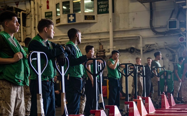 A line of military personnel wearing green safety vests are standing alongside a row of red manual pallet jacks inside what appears to be the hangar bay of a naval vessel. The personnel include both Marines in camouflage utilities and sailors in navy blue coveralls. They appear to be staged and ready for a cargo handling or resupply operation.