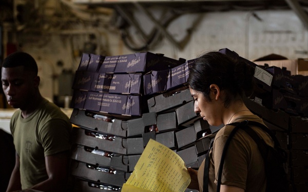 Two military personnel in olive/khaki uniforms are working in what appears to be a supply or storage area, likely aboard a naval vessel or military facility. A female service member is reviewing a yellow document/manifest while standing near stacked boxes and crates. Various food supply boxes are visible, including produce containers labeled 'The Grape' and a box marked 'CHILL', along with stacked dark mushroom product boxes in the background.
