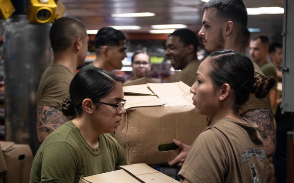 A group of military personnel, appearing to be U.S. Marines and Navy sailors, are engaged in a logistics or resupply operation in what appears to be a ship's storage compartment or supply room. Two female service members in the foreground are actively passing and handling cardboard boxes, while additional personnel form a working party line in the background. The setting suggests an underway replenishment or stores loading evolution aboard a naval vessel.