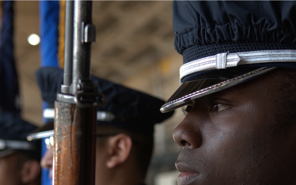 The image shows military or ceremonial honor guard members standing at attention in formal dress uniforms. The foreground subject is in sharp focus, holding a rifle with a wooden stock, wearing a dark blue dress uniform with a ceremonial cap featuring a silver band. Additional honor guard members are visible in the background, slightly out of focus, with at least one carrying a flag.
