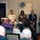 A panel discussion or conference event is taking place in what appears to be a hotel or conference room setting. Five panelists are seated at the front, including one individual in a white U.S. Navy officer uniform, while a moderator stands at a podium on the left. An audience is visible in the foreground, some with laptops open.