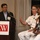Three individuals are participating in a panel discussion or presentation at an FCW (Federal Computer Week) event. A man in a dark suit stands at the podium with a microphone, while a military officer in white dress uniform and a woman in a floral dress are seated beside him. The military officer appears to be speaking and gesturing with his hand.