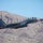 Red Flag 19-3 take offs A U.S. Air Force F-15E Strike Eagle jet fighter is captured in low-level flight against a backdrop of arid, rocky mountains under a clear blue sky. The aircraft displays distinctive markings including a red checkerboard nose pattern, 'LN' tail code, and serial number AF 97-219, indicating it belongs to a unit based at RAF Lakenheath, UK. The aircraft is fully loaded with underwing ordnance and appears to be flying at high speed through a desert canyon environment.