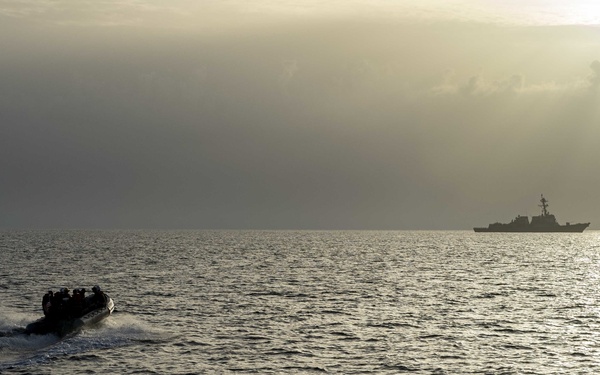A small rigid inflatable boat (RIB) carrying multiple personnel is moving at speed across open ocean water toward a larger naval warship visible on the horizon. The image is captured in low-light, backlit conditions with a hazy sky, giving the scene a dramatic silhouette effect. The composition suggests a military maritime operation, likely an insertion or transit exercise.