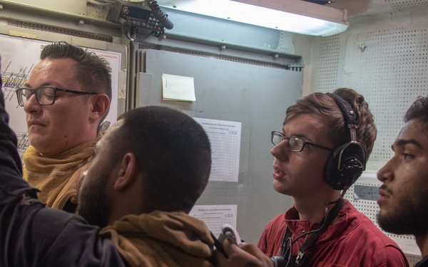 Four individuals are gathered in what appears to be a confined naval workspace or control room aboard a U.S. Navy vessel. One person wearing a red shirt with 'U.S. Navy' markings and headset appears to be communicating, while others examine documents or charts on a wall-mounted board. The setting suggests a training or operational briefing scenario.