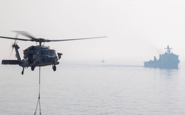 A military helicopter, appearing to be an MH-60 Seahawk, is conducting a vertical replenishment (VERTREP) operation over the deck of a naval vessel. The helicopter is lowering or retrieving cargo nets filled with supplies onto the ship's flight deck. At least two additional naval vessels are visible in the background on calm, hazy seas.