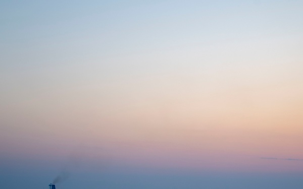 A large ship is silhouetted against a calm sea during twilight, either at dawn or dusk. The sky displays a gradient of soft pastel colors ranging from blue-gray at the top to warm orange and pink near the horizon. The water is nearly flat with gentle ripples reflecting the ambient light.
