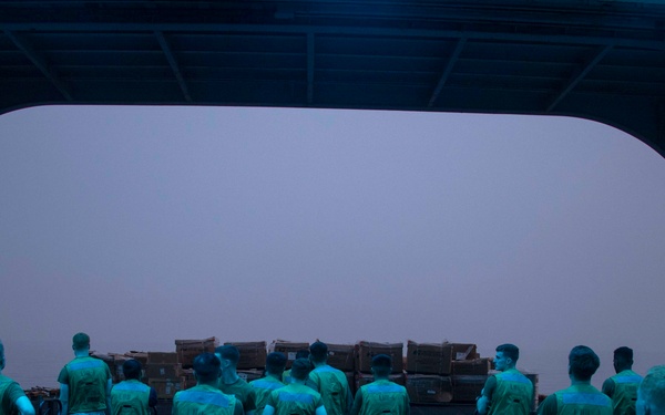 A group of military personnel, likely U.S. Marines based on their camouflage uniforms and green t-shirts, are gathered inside what appears to be the cargo bay or well deck of a naval vessel. They are positioned near pallet jacks facing a large open bay door revealing an overcast sky, with stacked wooden crates/cargo visible near the opening. The scene suggests a logistics or cargo offloading operation at dawn or dusk.