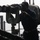 A person in dark blue uniform is using large mounted binoculars or a telescope on what appears to be the deck of a naval vessel. The figure is silhouetted against a hazy, overcast sky, suggesting maritime surveillance or observation duties. Various covered equipment and metal structures are visible in the background.