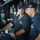 Two U.S. Navy sailors are stationed at the helm/bridge of USS Sentry (MCM 3), a mine countermeasures ship. The sailor on the left is operating the ship's wheel while the one on the right observes, both wearing Navy working uniforms and ship-specific ball caps. The setting is the interior bridge/pilothouse with various navigation and communication equipment visible.
