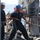 Two naval personnel are working on the deck of a military vessel at sea. One sailor wearing a blue hard hat is actively pulling a thick rope or line, while a second sailor in a white hard hat observes. The scene depicts routine deck operations or mooring/line-handling procedures aboard a warship.