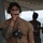 A military service member wearing a tan/coyote brown t-shirt and tactical headset is speaking into a handheld microphone or push-to-talk device. He has a prominent Polynesian-style tribal tattoo on his left arm and is wearing a dark baseball cap. Two other individuals are visible in the blurred background, one wearing a garment with 'NAVY' text visible.