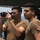 Two individuals wearing matching tan/coyote brown military-style t-shirts are standing on what appears to be a naval vessel deck. One person is holding and operating a Sony HD handheld camcorder, while the other stands nearby observing. A third person is partially visible in the background, and large binocular equipment is visible on the left side of the frame.