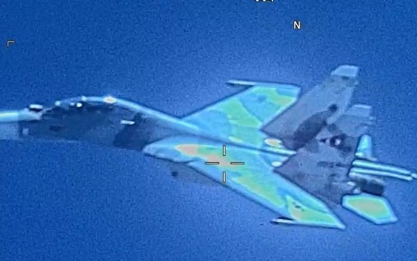 An aerial surveillance or targeting camera image showing a military jet fighter aircraft in flight against a blue sky background. The aircraft displays a camouflage paint scheme with green, tan, and dark tones, and appears to be carrying underwing ordnance or fuel tanks. A targeting reticle crosshair is visible overlaid on the aircraft, along with a compass indicator showing north at the top of the frame.